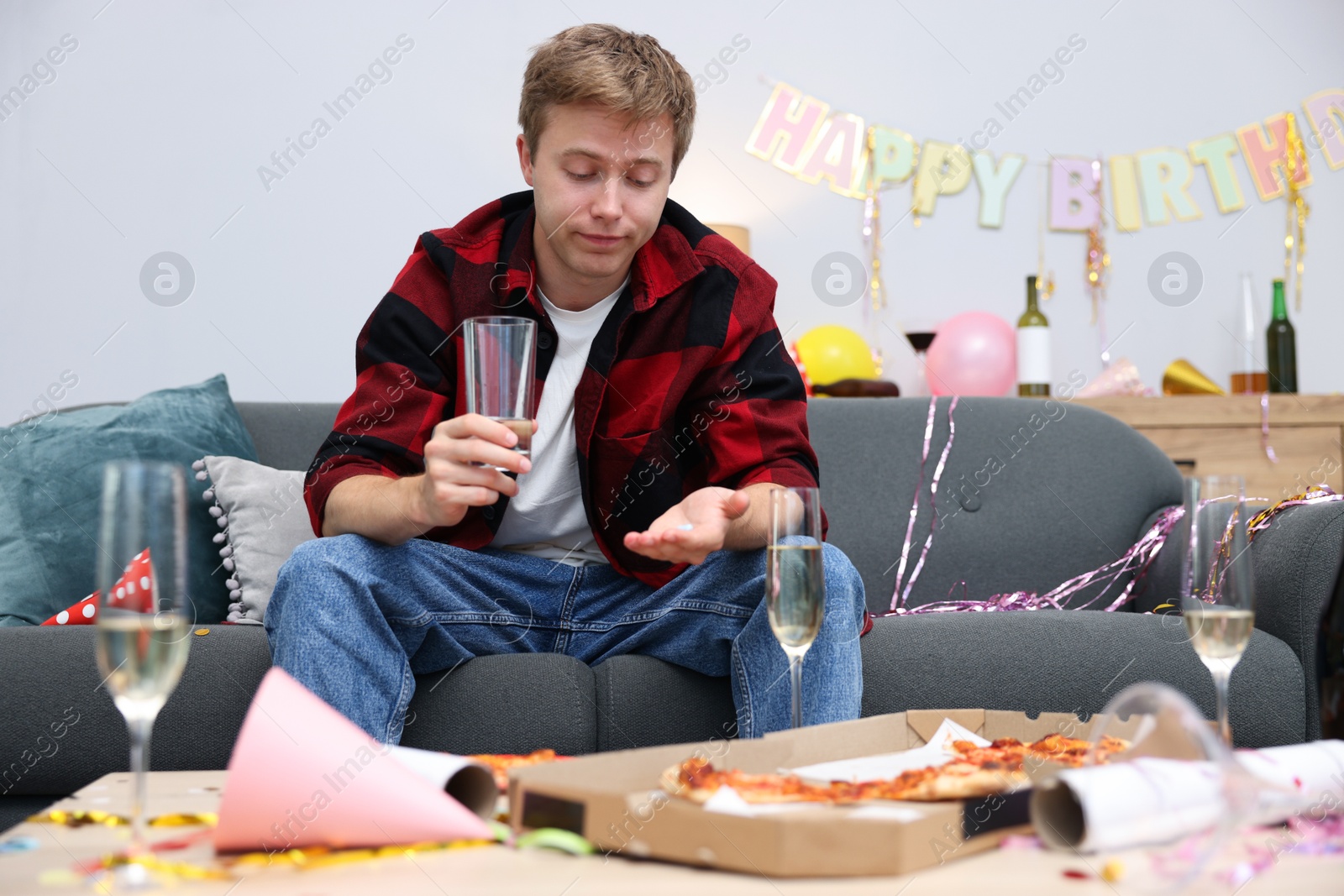 Man with glass of water and pills suffering from hangover on sofa in messy room after party Photo of Man with glass of water and pills suffering from hangover on sofa in messy room after party