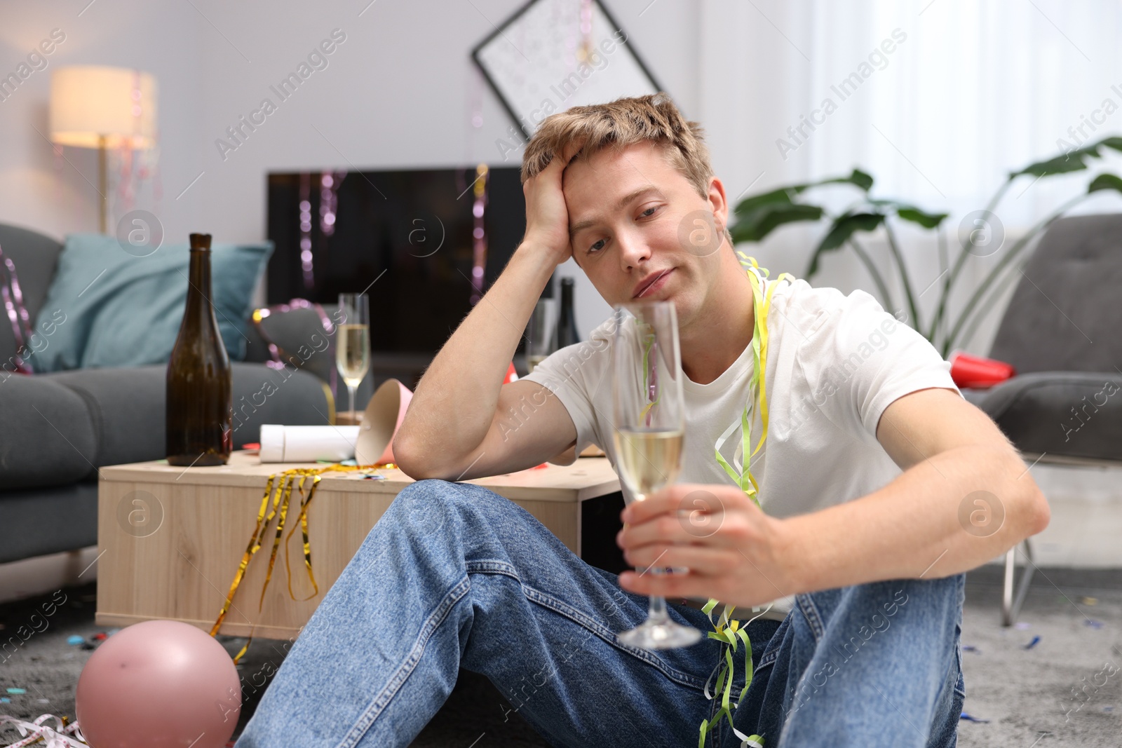 Man with glass of alcohol suffering from hangover on floor in messy room after party Photo of Man with glass of alcohol suffering from hangover on floor in messy room after party
