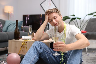 Photo of Man with glass of alcohol suffering from hangover on floor in messy room after party