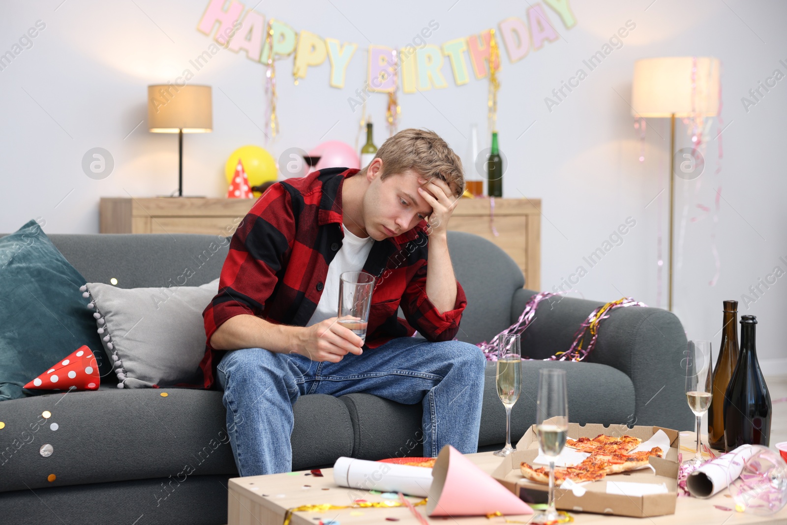 Man with glass of water suffering from hangover on sofa in messy room after party Photo of Man with glass of water suffering from hangover on sofa in messy room after party