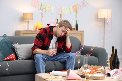 Photo of Man with glass of water suffering from hangover on sofa in messy room after party