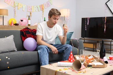 Photo of Man with glass of alcohol suffering from hangover on sofa in messy room after party