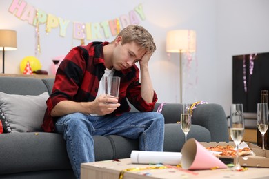 Photo of Man with glass of water suffering from hangover on sofa in messy room after party