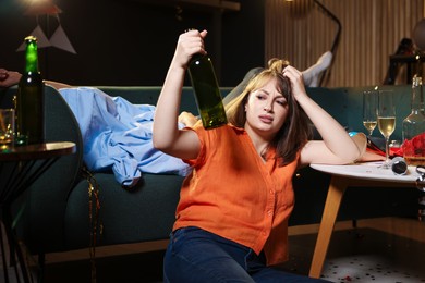 Photo of Woman with bottle of alcohol suffering from hangover in messy room after party