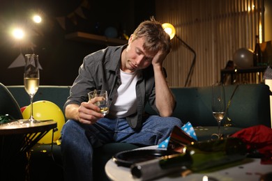Photo of Man with glass of alcohol suffering from hangover on sofa in messy room after party