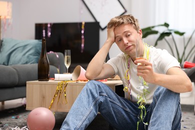 Photo of Man with glass of alcohol suffering from hangover on floor in messy room after party