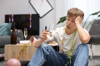 Photo of Man with glass of alcohol suffering from hangover on floor in messy room after party