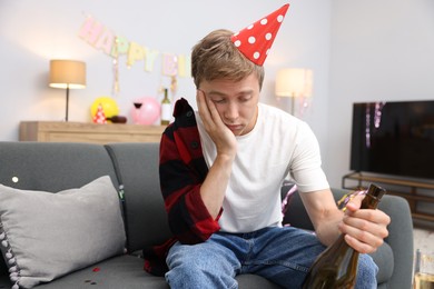 Photo of Man with bottle of alcohol suffering from hangover on sofa in messy room after party