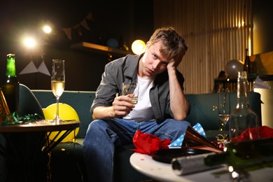 Photo of Man with glass of alcohol suffering from hangover on sofa in messy room after party