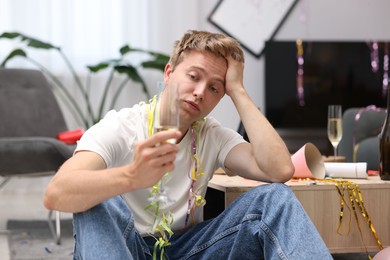 Photo of Man with glass of alcohol suffering from hangover on floor in messy room after party