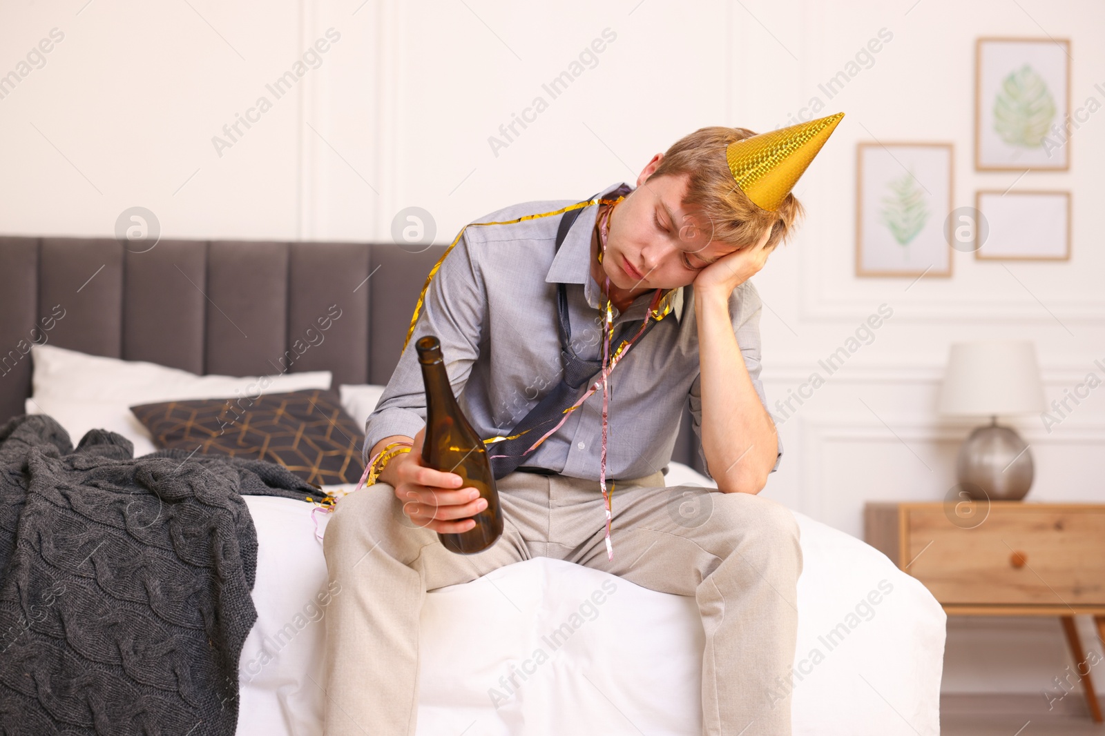 Man with bottle of alcohol suffering from hangover on bed in messy room after party Photo of Man with bottle of alcohol suffering from hangover on bed in messy room after party