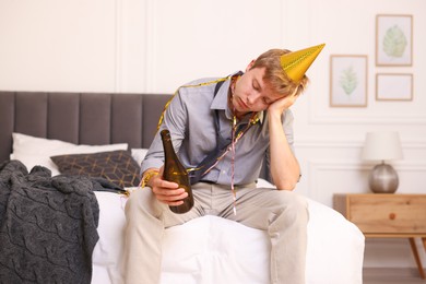 Photo of Man with bottle of alcohol suffering from hangover on bed in messy room after party