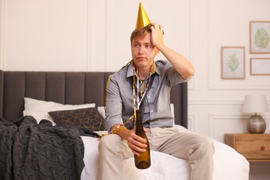 Photo of Man with bottle of alcohol suffering from hangover on bed in messy room after party