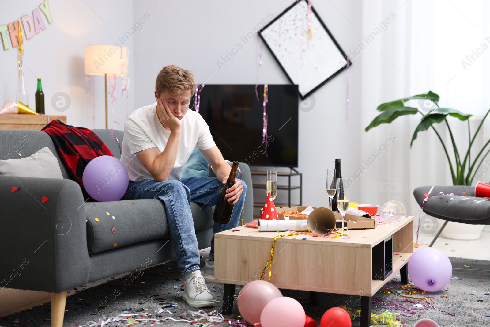 Man with bottle of alcohol suffering from hangover on sofa in messy room after party Photo of Man with bottle of alcohol suffering from hangover on sofa in messy room after party