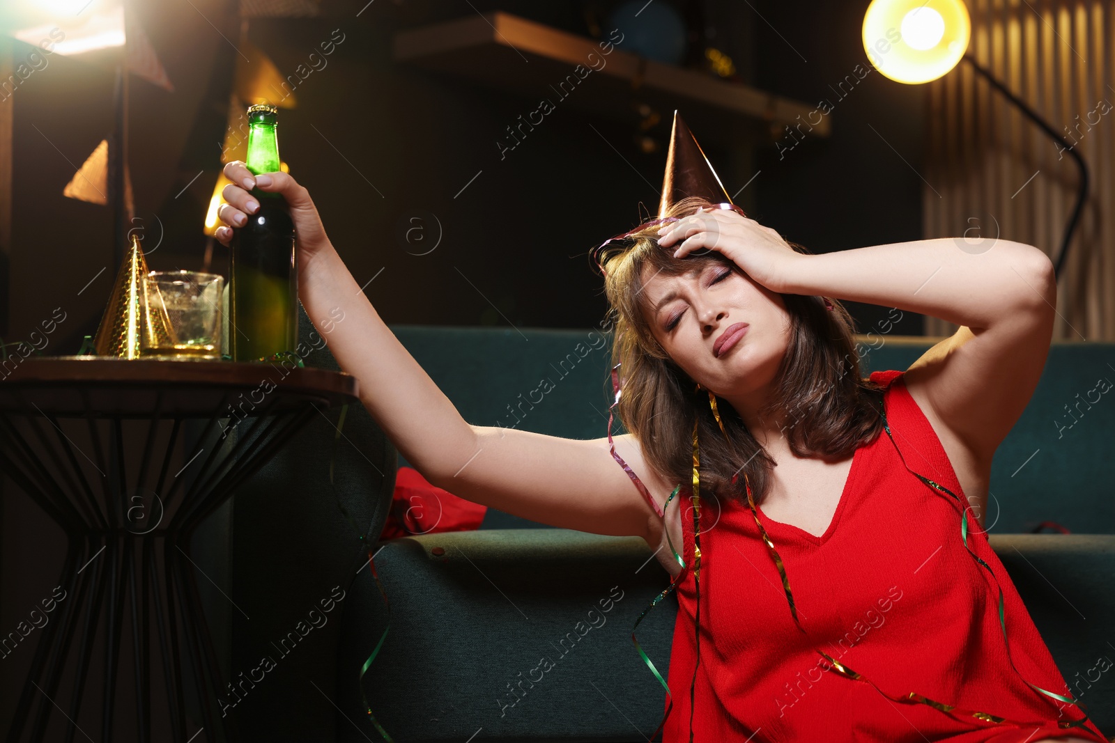 Photo of Woman with bottle of beer suffering from hangover in messy room after party