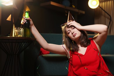 Photo of Woman with bottle of beer suffering from hangover in messy room after party