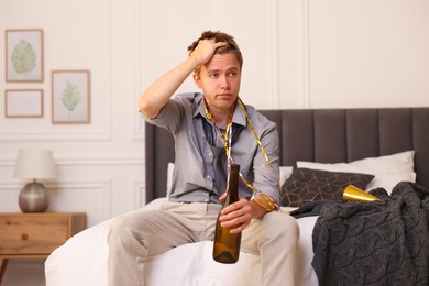 Photo of Man with bottle of alcohol suffering from hangover on bed in messy room after party