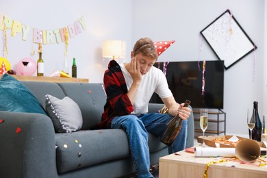 Photo of Man with bottle of alcohol suffering from hangover on sofa in messy room after party
