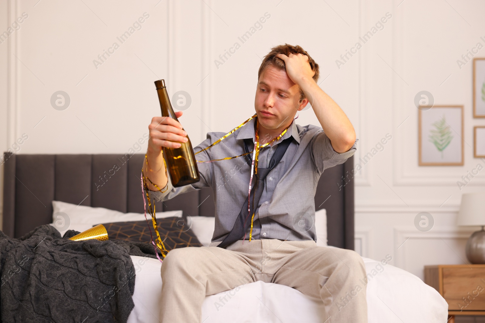 Man with bottle of alcohol suffering from hangover on bed in messy room after party Photo of Man with bottle of alcohol suffering from hangover on bed in messy room after party