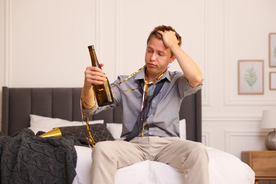Photo of Man with bottle of alcohol suffering from hangover on bed in messy room after party