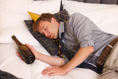 Photo of Man with bottle of alcohol suffering from hangover on bed in messy room after party