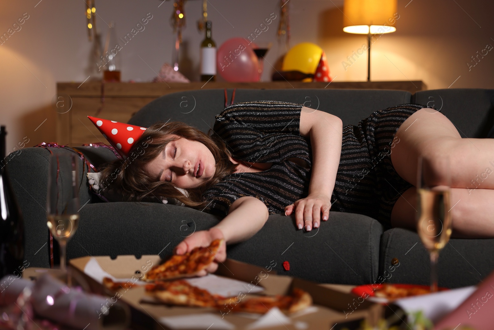 Woman with piece of pizza suffering from hangover on sofa in messy room after party Photo of Woman with piece of pizza suffering from hangover on sofa in messy room after party