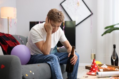 Photo of Man with bottle of alcohol suffering from hangover on sofa in messy room after party