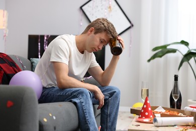 Photo of Man with bottle of alcohol suffering from hangover on sofa in messy room after party