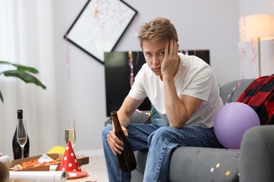 Photo of Man with bottle of alcohol suffering from hangover on sofa in messy room after party