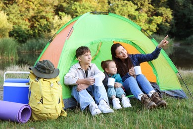 Photo of Happy family spending time together with camping tent, tourist equipment and mugs of hot drink outdoors