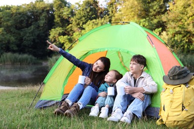 Photo of Happy family spending time together with camping tent, tourist equipment and mugs of hot drink outdoors