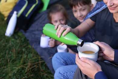 Photo of Mother pouring drink from bottle into mug and her children outdoors, closeup