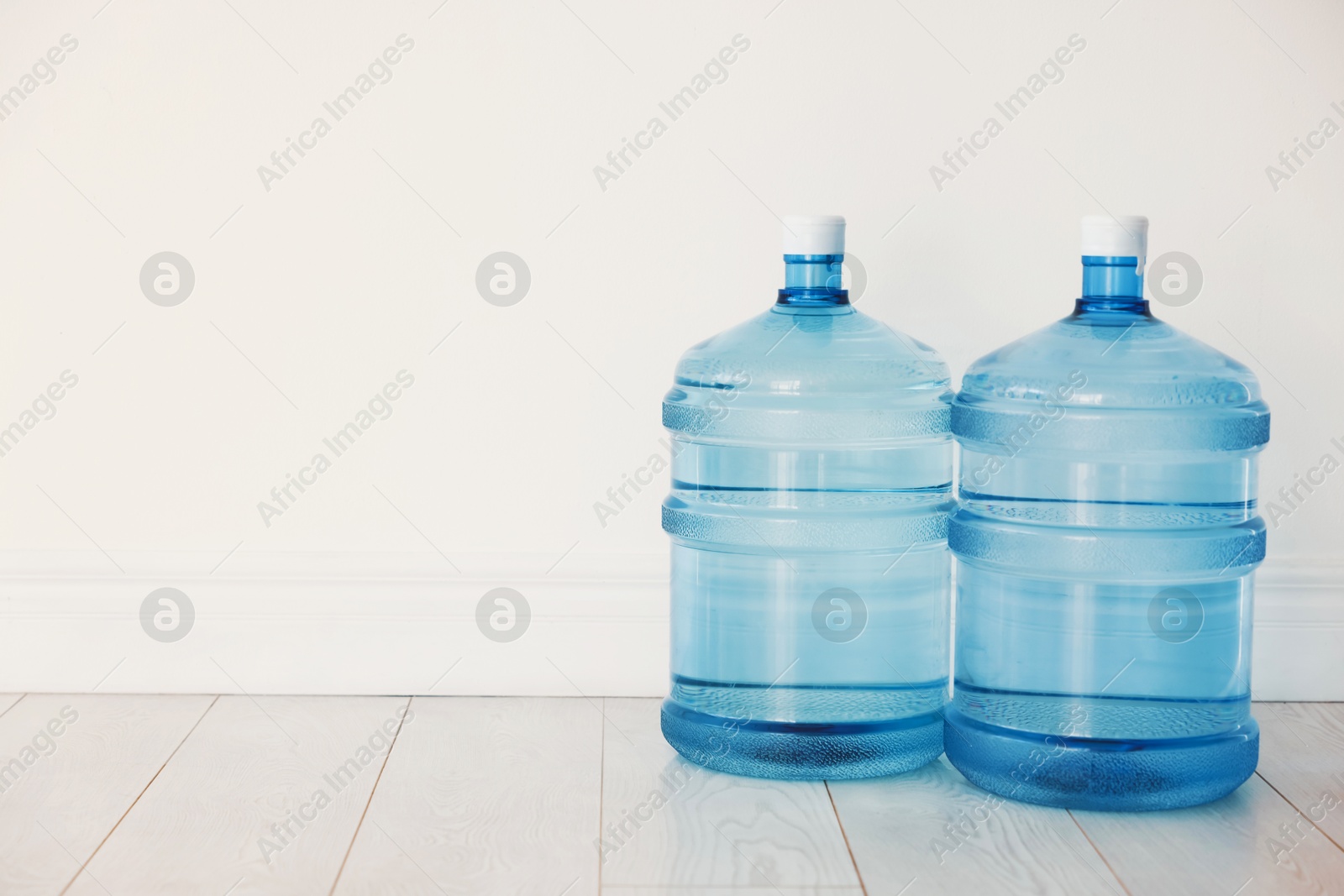 Cooler bottles with water on wooden floor near white wall. Space for text Photo of Cooler bottles with water on wooden floor near white wall. Space for text