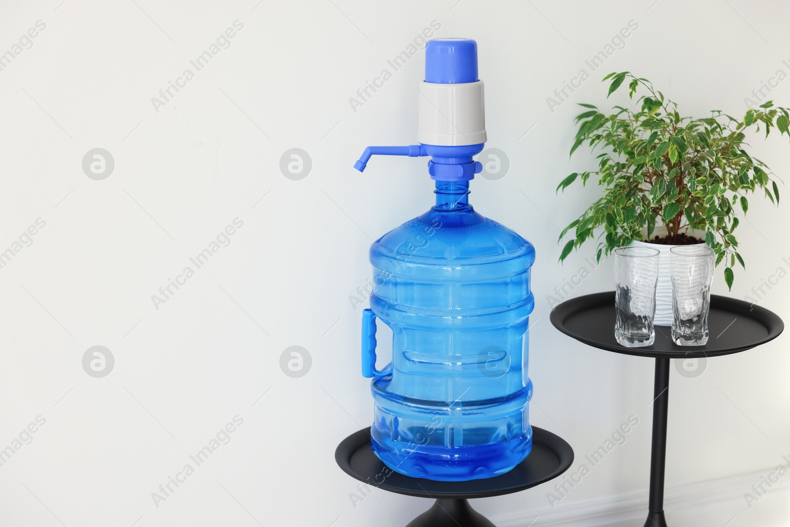 Cooler bottle with water, pump, glasses and houseplant near white wall. Space for text Photo of Cooler bottle with water, pump, glasses and houseplant near white wall. Space for text