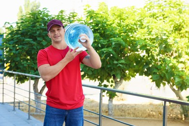Photo of Smiling delivery man with cooler bottle of water near railings outdoors. Space for text