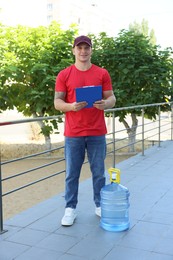 Photo of Smiling delivery man with cooler bottle of water and clipboard near railings outdoors