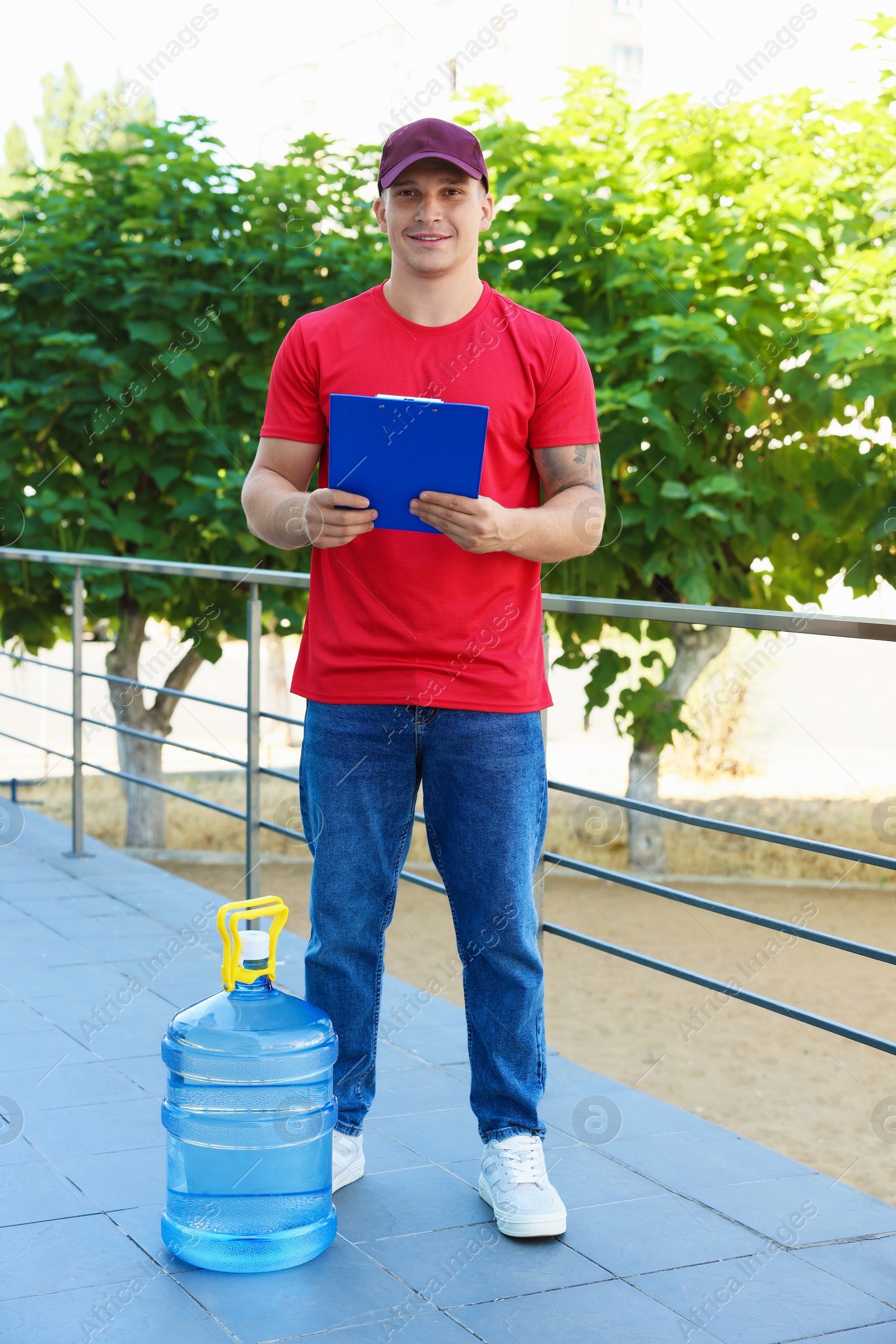 Photo of Smiling delivery man with cooler bottle of water and clipboard near railings outdoors
