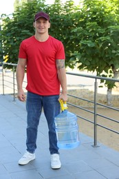 Photo of Smiling delivery man with cooler bottle of water near railings outdoors