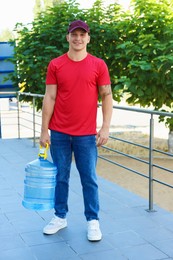 Photo of Smiling delivery man with cooler bottle of water near railings outdoors