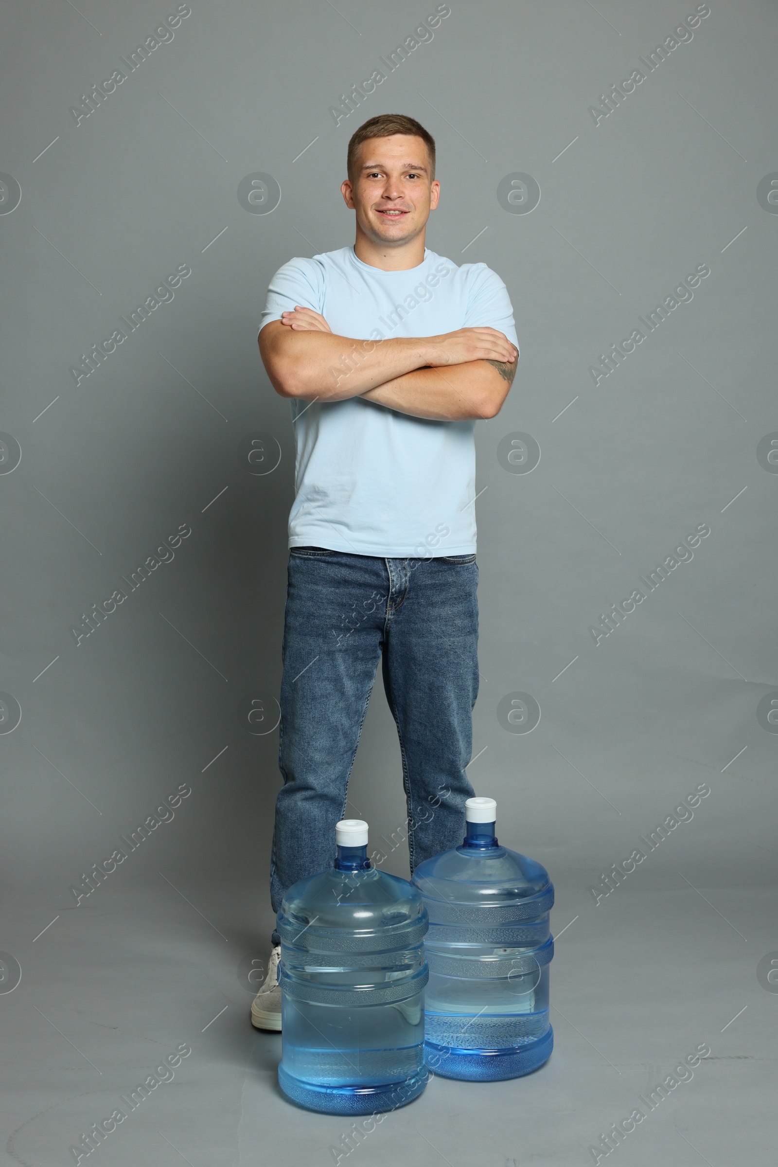 Photo of Smiling delivery man with crossed arms and cooler bottles of water on grey background