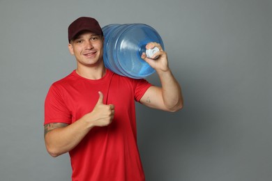 Photo of Smiling delivery man with cooler bottle of water showing thumbs up on grey background. Space for text