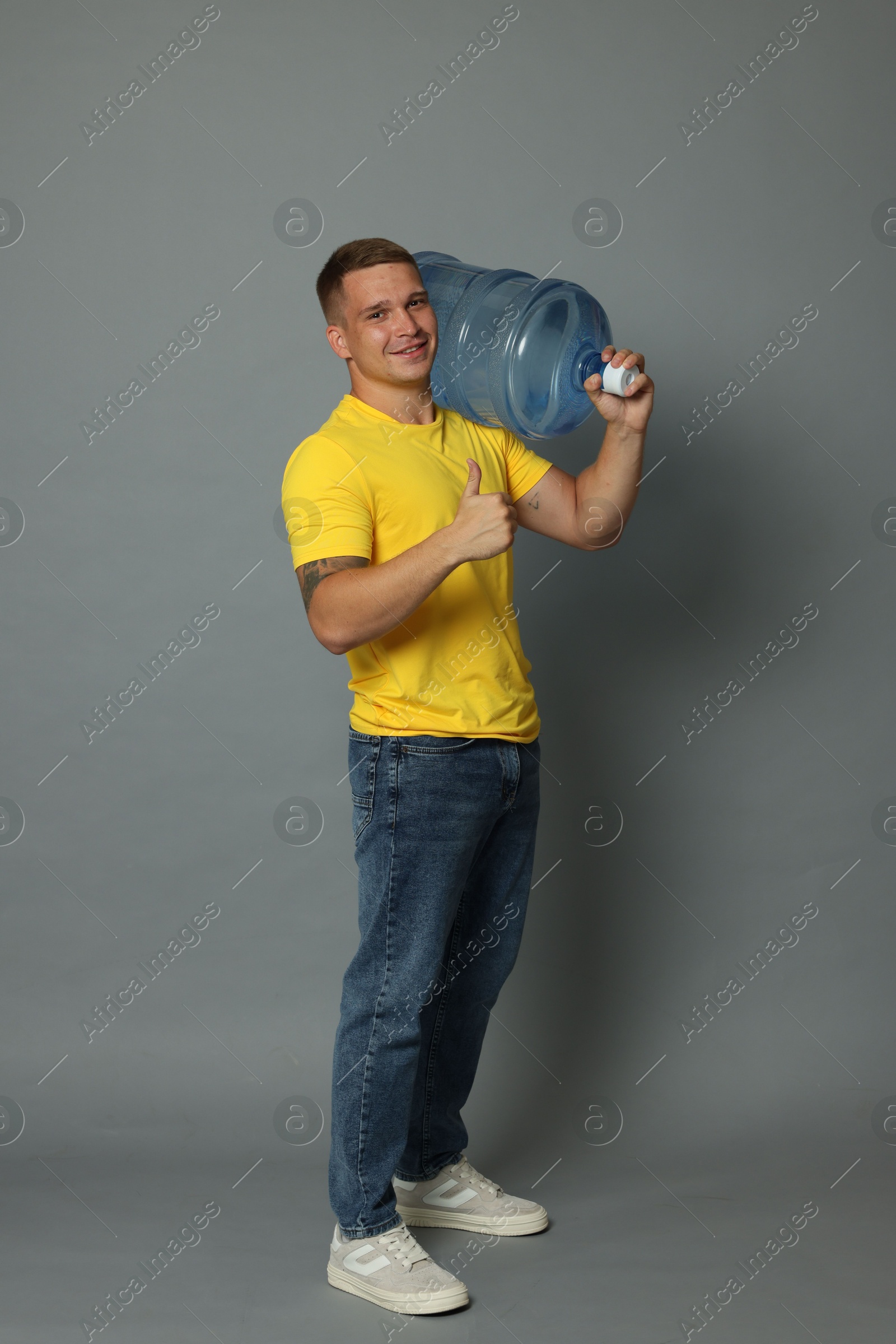 Photo of Smiling delivery man with cooler bottle of water showing thumbs up on grey background