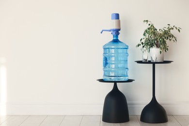 Photo of Cooler bottle with water, pump, glasses and houseplant near white wall. Space for text