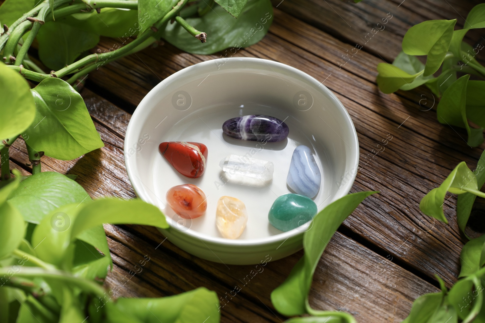 Photo of Aura. Different natural mineral stones in bowl and green leaves on wooden table, closeup