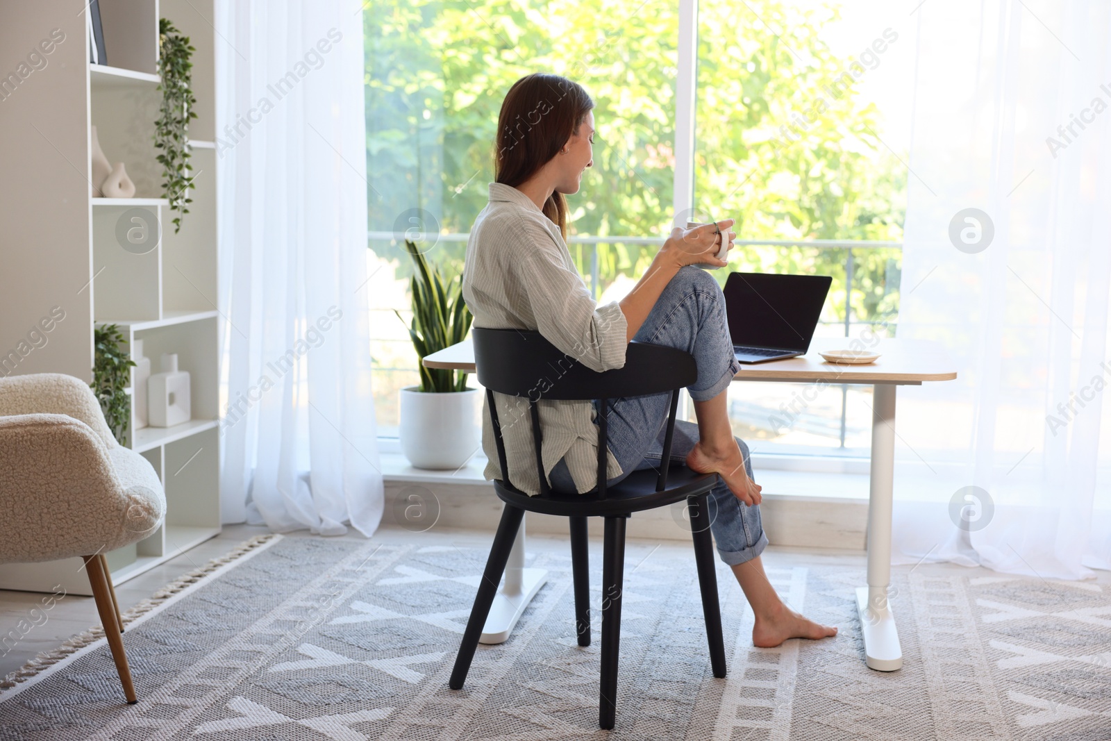 Photo of Woman with coffee at wooden desk indoors, back view. Home workplace