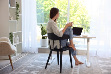 Photo of Woman with coffee at wooden desk indoors, back view. Home workplace