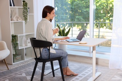 Photo of Woman with coffee using laptop at wooden desk indoors. Home workplace