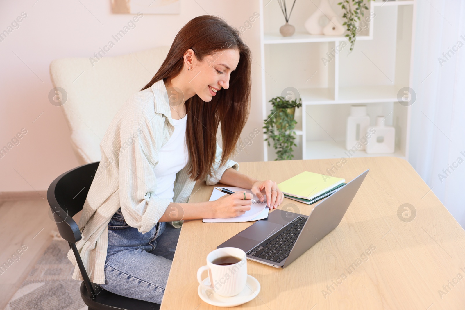Photo of Woman writing and using laptop at wooden desk indoors. Home workplace