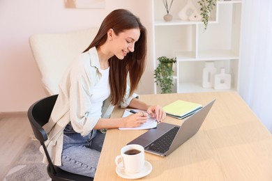Photo of Woman writing and using laptop at wooden desk indoors. Home workplace