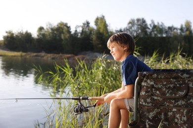 Photo of Little boy sitting on folding chair and fishing near river outdoors
